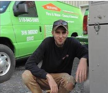 A photo of a smiling male employee wearing a black SERVPRO shirt crouching in front of a green SERVPRO van.