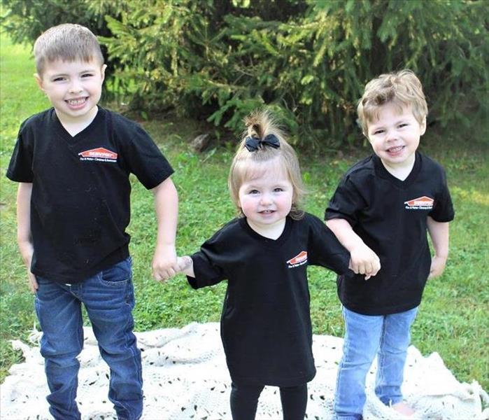 3 smiling children standing in a line holding hands, smiling and wearing black tshirts with the word SERVPRO on it.