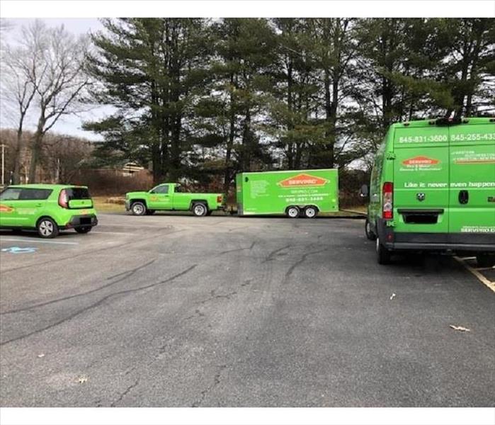 3 SERVPRO vehicles and a tow-behind trailer in a parking lot in front of a commercial building.