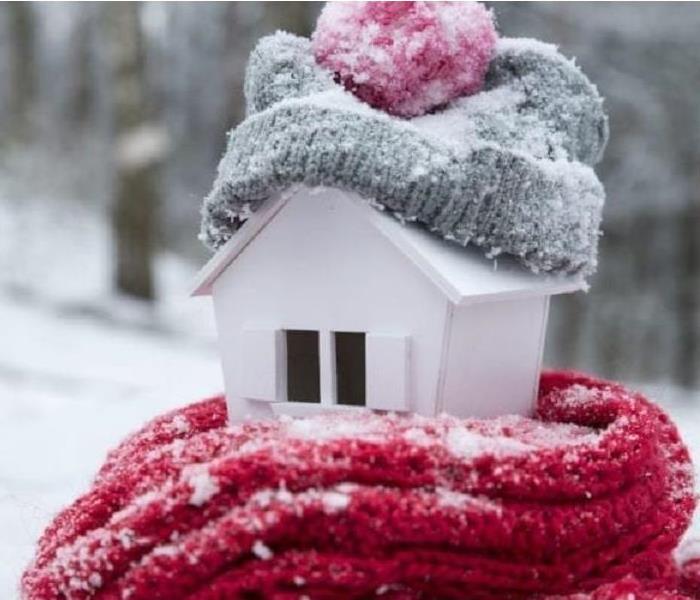 An image of a little white bird house with a red scarf wrapped around the base and a grey winter hat on top, covered in snow.