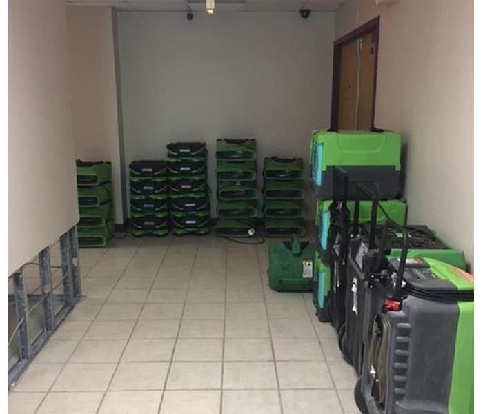 SERVPRO dehumidifiers and air movers lined up against a wall in a commercial building after water damage remediation