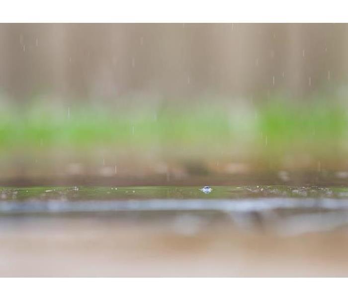 A close up of raindrops falling on a concrete sidewalk with green grass in the background.