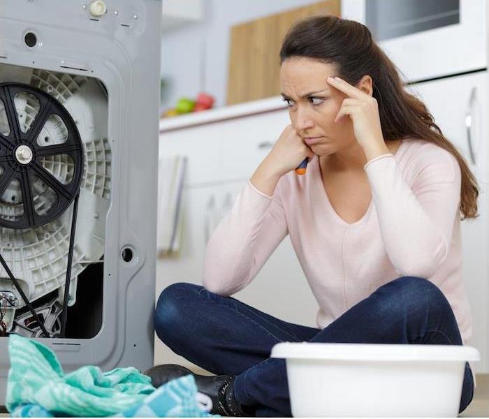 A woman sitting on the floor looking frustrated next to a broken washing machine