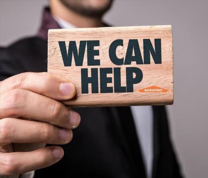 A man in a jacket and tie holding up a small wooden block with the words "We Can Help" printed on it in black ink.