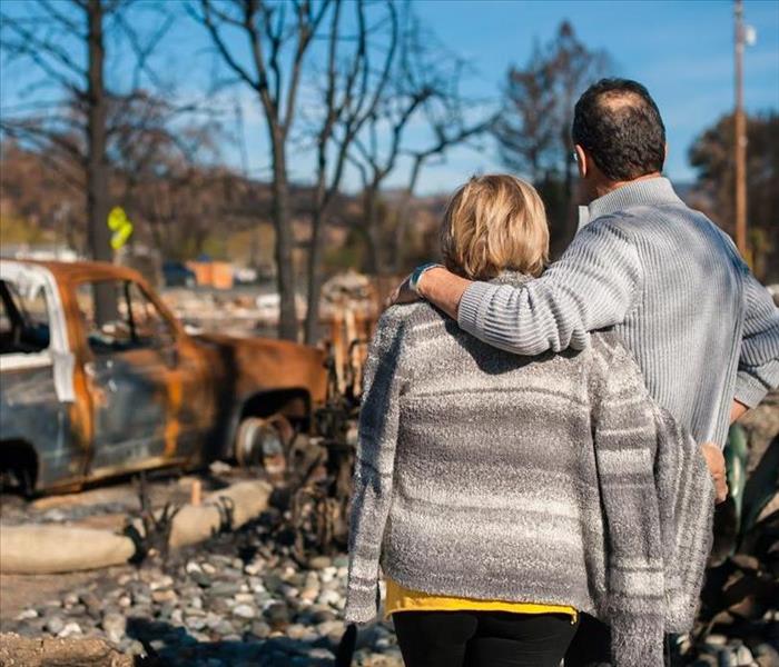 A couple comforting each other while looking at the remains of fire damage to their home and vehicle